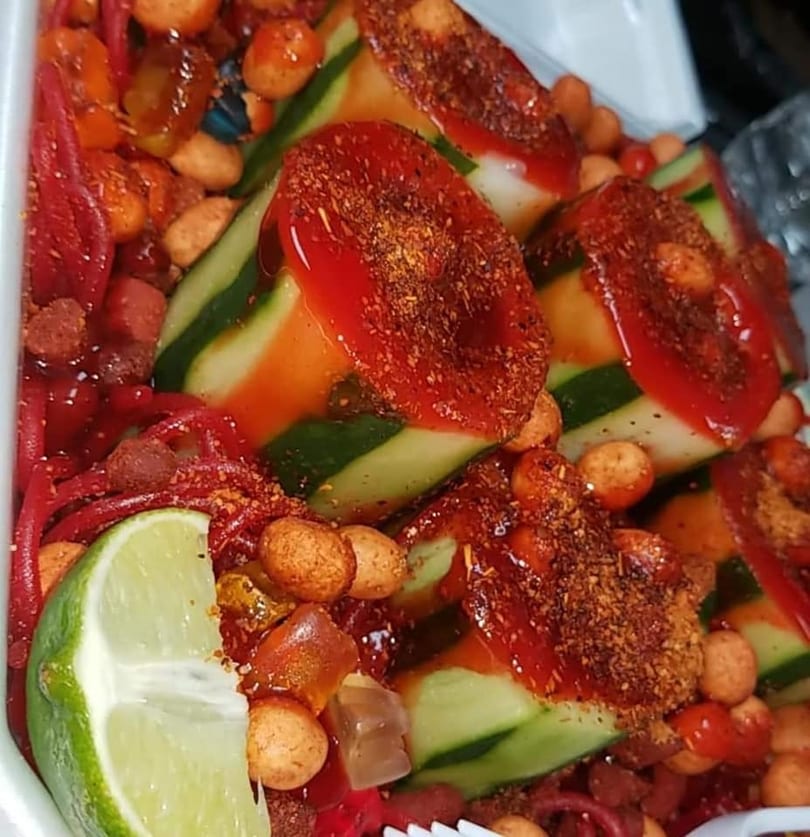 Spicy cucumber slices with chili powder, hot sauce, peanuts, and a lime wedge.