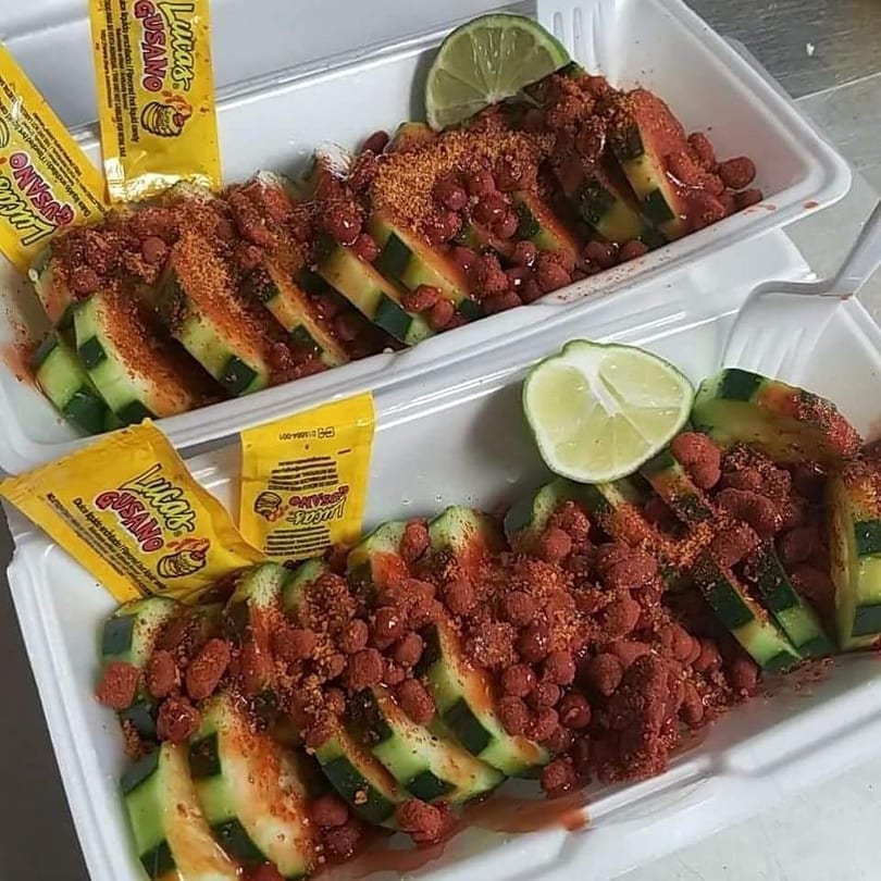 Two servings of sliced cucumbers topped with red candy, chili powder, and lime wedges.