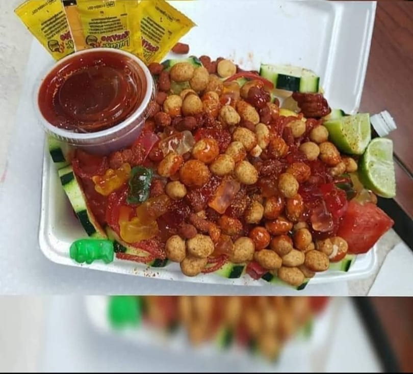 Mexican snack with peanuts, gummy bears, cucumber, watermelon, lime, and chili sauce in a to-go container.