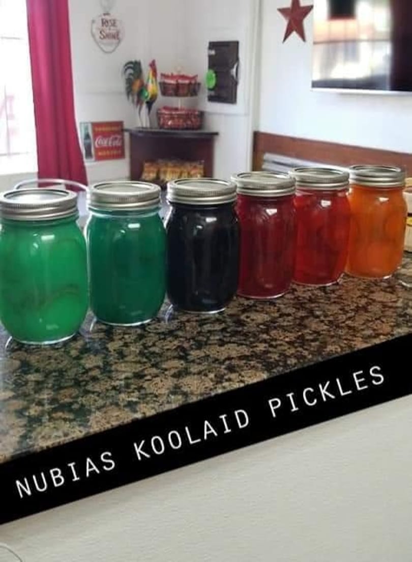 Six colorful jars of Kool-Aid pickles, including green, black, red, and orange, on a speckled countertop.