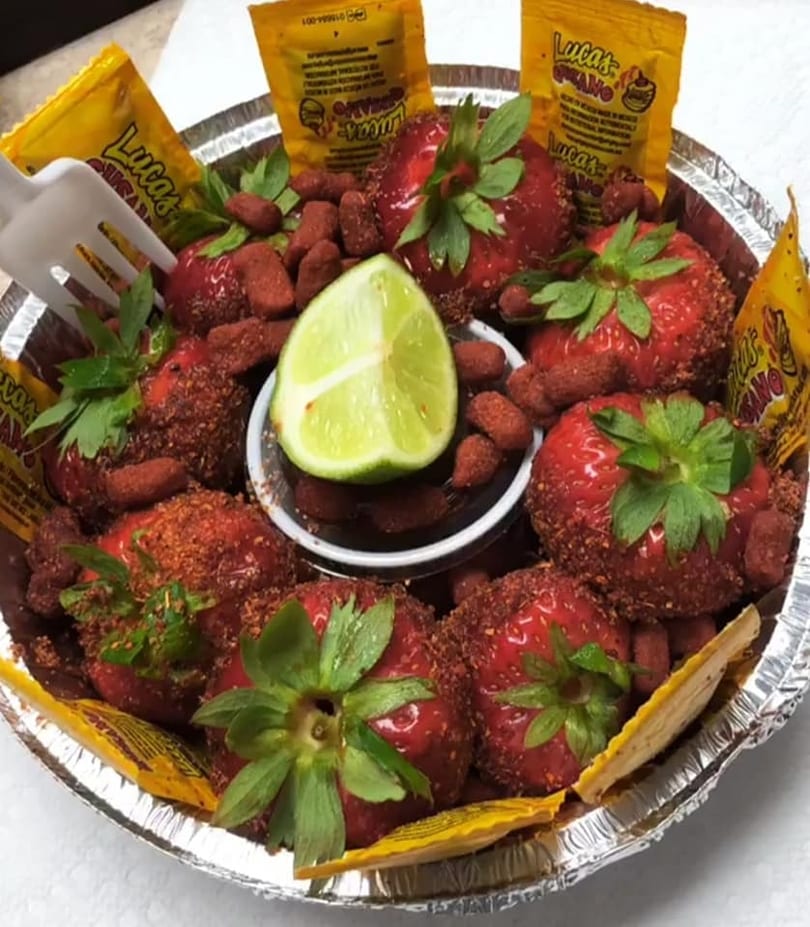 Strawberries coated in chili powder, with a lime wedge and Lucas candy packets in a foil tray.